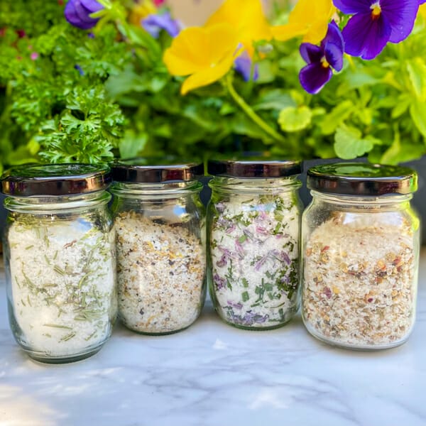 Four finishing salts with fresh herbs growing in the background. Salts include rosemary, lemon pepper, chive and chili lime finishing salts.