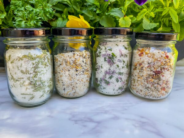 Four finishing salts with fresh herbs growing in the background. Salts include rosemary, lemon pepper, chive and chili lime finishing salts.