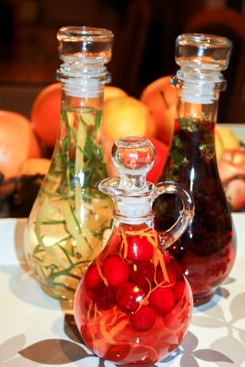 3 bottles of infused vinegar with fruits arranged in the background.