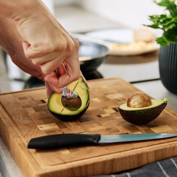 hand with spoon scooping out avocado resting on cutting board next to knife