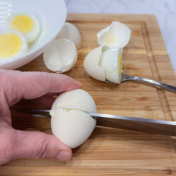 large knife through middle of hard cooked egg on board with other egg shells and spoon in cut egg