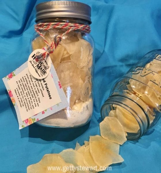 Jar of scalloped potatoes with gift tag, instructions and decorative ribbon. Dry potato slices spread out beside the jar.