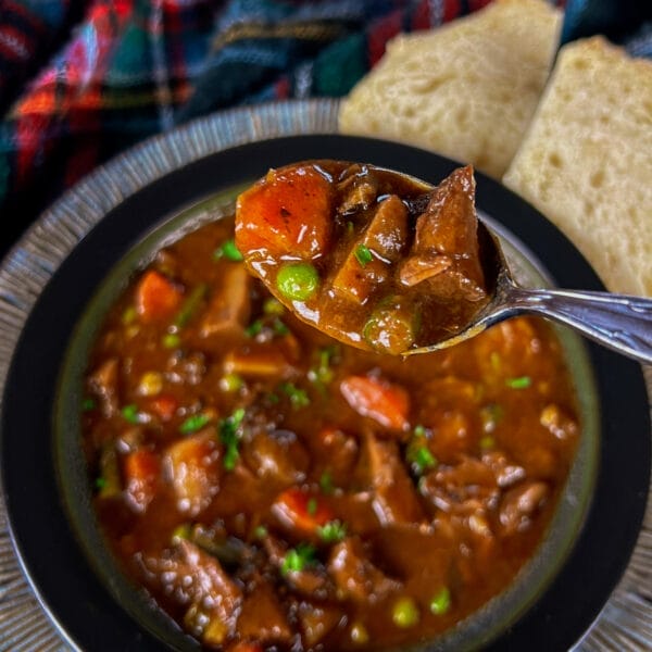 spoon of beef stew over bowl of beef stew on big plate with side of bread