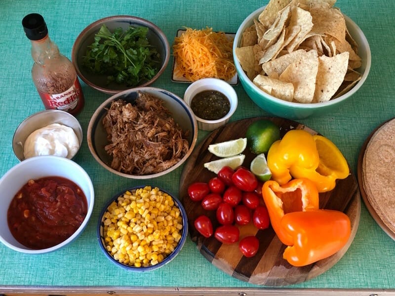 pulled pork tacos fixings: cilantro, cheese, veggies, corn, salsa, sour cream, tortilla wraps and chips