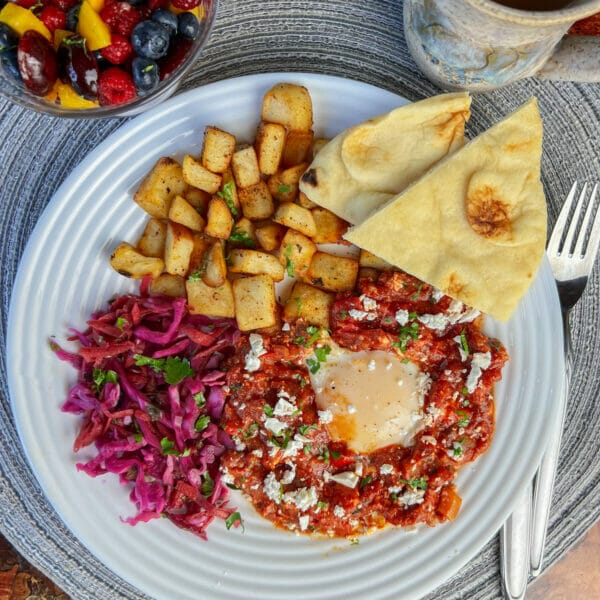 shakshuka egg on plate with red cabbage, roasted potatoes, naan and fruit salad on the side