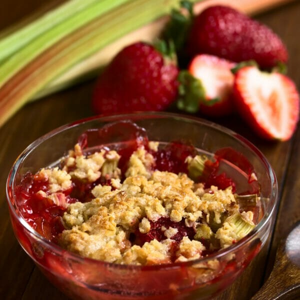 a close up image of a small bowl of strawberry rhubarb crisp. Strawberries and rhubarb are seen in the background.