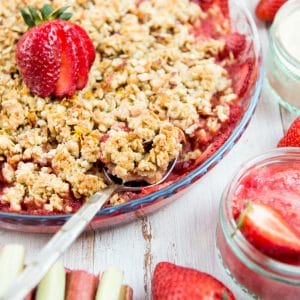 Bright red strawberry sits on top of strawberry rhubarb crisp. Rhubarb and strawberries surround.