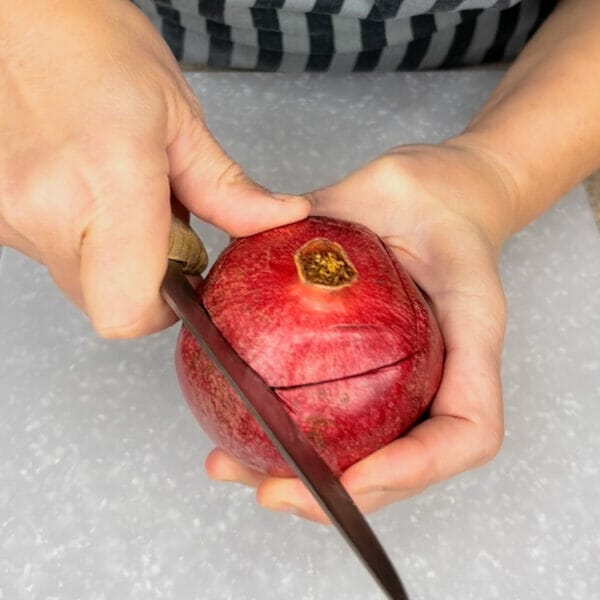 hands cutting top of pomegranate with knife over cutting board