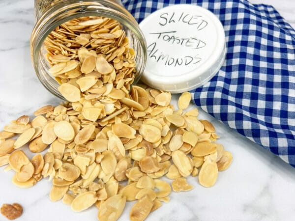 sliced toasted almonds spilling out of jar with lid and blue checked cloth
