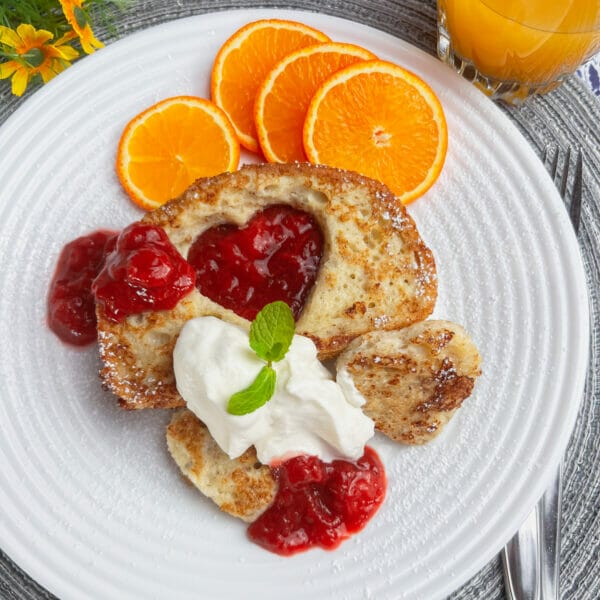 French toast with heart cutouts, red fruit sauce, oranges and whipped cream.