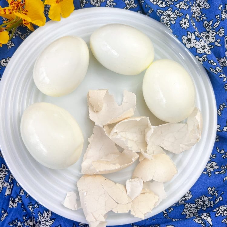 4 boiled and peeled eggs on a plate with shells beside, with blue tablecloth in the background