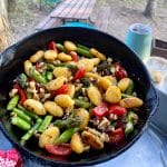 cast iron pan filled with asparagus and tomato gnocchi with picnic table and camping cup in background